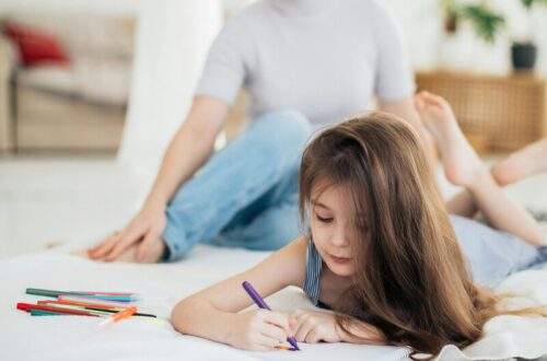 mãe, rosto desfocado, semblante sério, olha de longe sua filha que está deitada desenhando.
