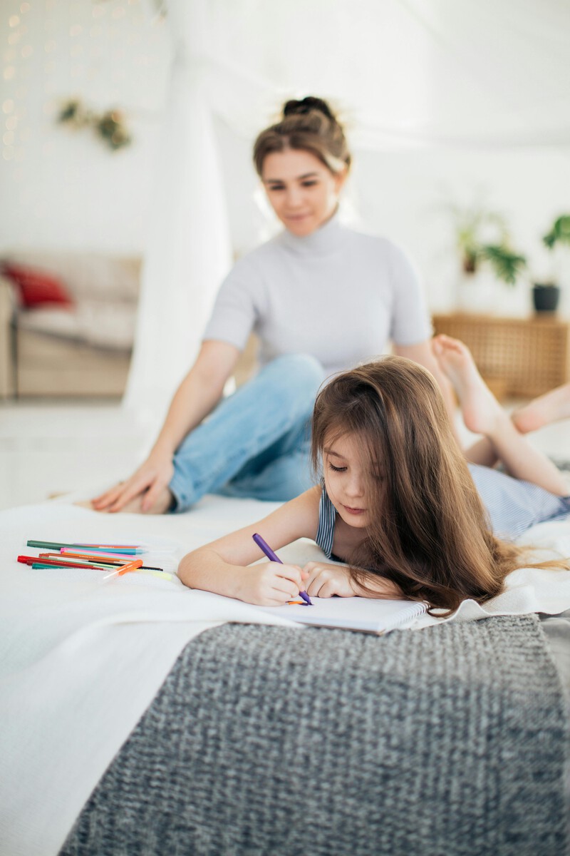 mãe, rosto desfocado, semblante sério, olha de longe sua filha que está deitada desenhando.