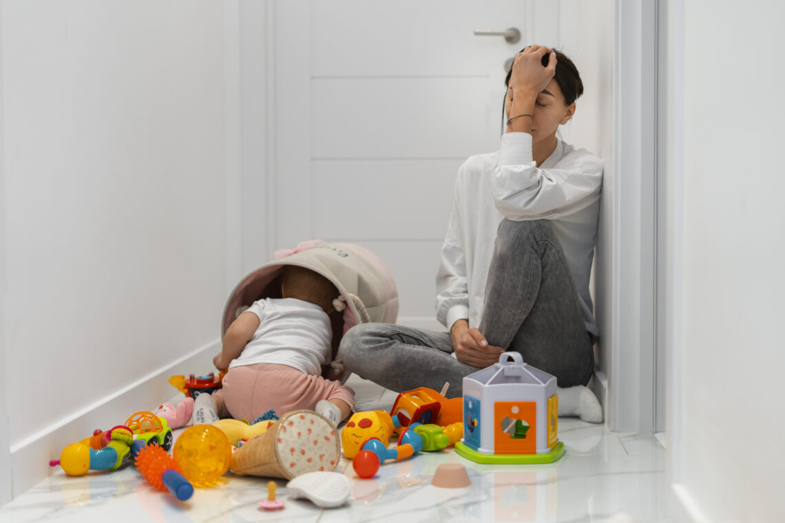 mãe exausta, sentada no chão com a crinaça brincando e brinquedos espalhados pelo chão